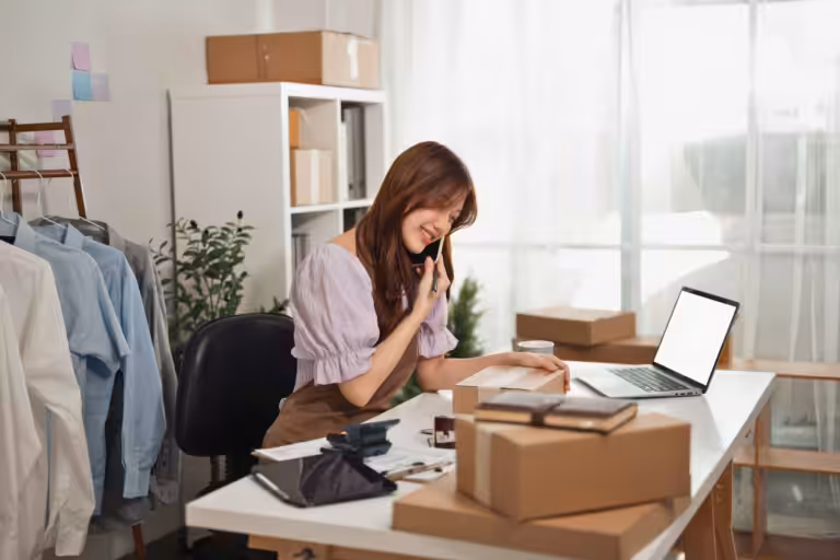Woman on the phone at a home office desk with a laptop, shipping boxes and a clothing rack, illustrating home-based business insurance